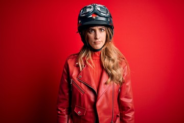 Young beautiful brunette motrocyclist woman wearing moto helmet over red background skeptic and...
