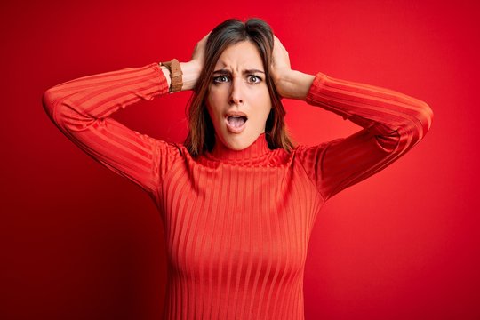 Young beautiful brunette woman wearing casual turtleneck sweater over red background Crazy and scared with hands on head, afraid and surprised of shock with open mouth