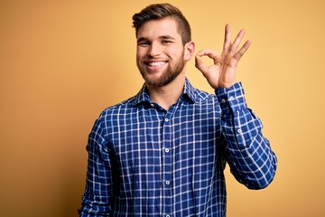 Fototapeta premium Young blond businessman with beard and blue eyes wearing shirt over yellow background smiling positive doing ok sign with hand and fingers. Successful expression.