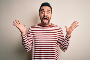 Naklejka premium Young handsome man with beard wearing casual striped t-shirt standing over white background celebrating crazy and amazed for success with arms raised and open eyes screaming excited. Winner concept
