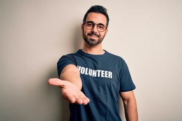 Handsome man with beard wearing t-shirt with volunteer message over white background smiling cheerful offering palm hand giving assistance and acceptance.