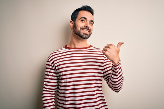 Young handsome man with beard wearing casual striped t-shirt standing over white background smiling with happy face looking and pointing to the side with thumb up.