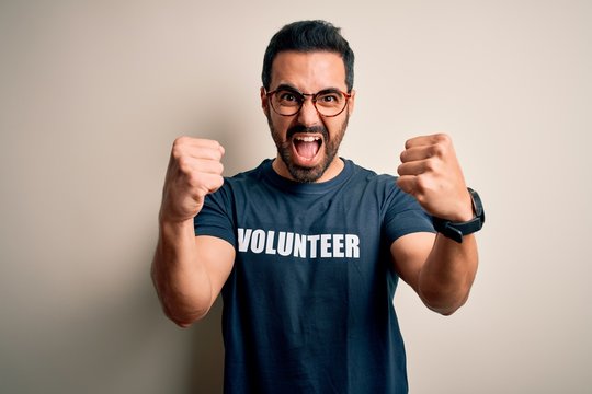 Handsome man with beard wearing t-shirt with volunteer message over white background angry and mad raising fists frustrated and furious while shouting with anger. Rage and aggressive concept.
