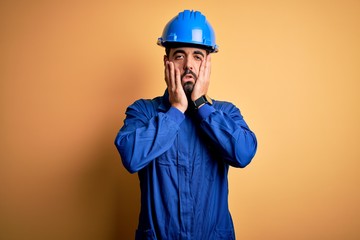 Mechanic man with beard wearing blue uniform and safety helmet over yellow background Tired hands...