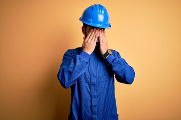 Mechanic man with beard wearing blue uniform and safety helmet over yellow background rubbing eyes...