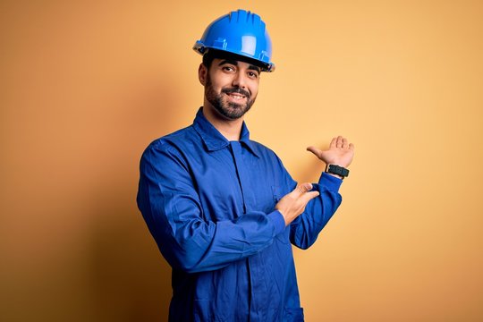 Mechanic man with beard wearing blue uniform and safety helmet over yellow background Inviting to enter smiling natural with open hand