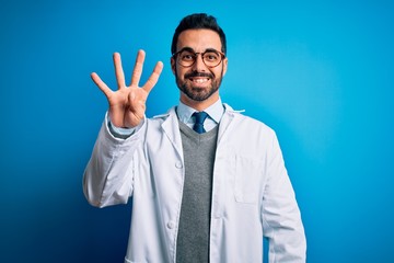 Young handsome doctor man with beard wearing coat and glasses over blue background showing and pointing up with fingers number four while smiling confident and happy.