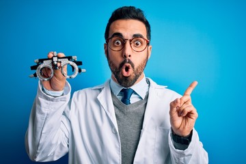 Young handsome optical man with beard holding optometry glasses over blue background Surprised pointing with finger to the side, open mouth amazed expression. - obrazy, fototapety, plakaty