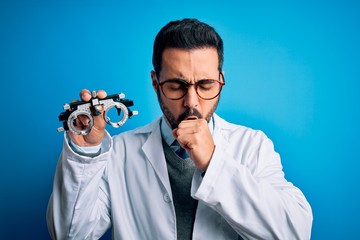 Young handsome optical man with beard holding optometry glasses over blue background feeling unwell...