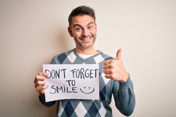 Young handsome man holding banner with funny positive message over white background happy with big smile doing ok sign, thumb up with fingers, excellent sign