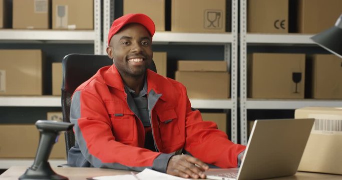 Young cheerful African American postman in red iniform and cap sitting at desk in postal office store and working at computer. Portrait of male courier smiling happily to camera in storage of parcels.