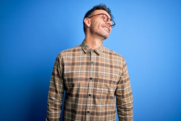 Young handsome man wearing casual shirt and glasses standing over blue background looking away to side with smile on face, natural expression. Laughing confident.