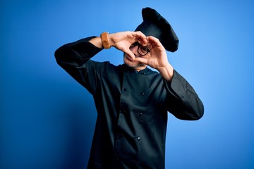 Young handsome chef man wearing cooker uniform and hat over isolated blue background Doing heart shape with hand and fingers smiling looking through sign