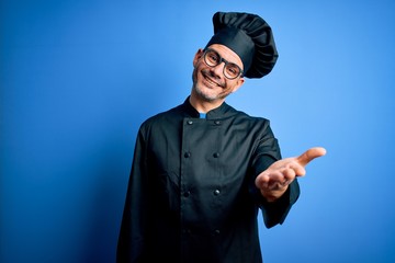 Young handsome chef man wearing cooker uniform and hat over isolated blue background smiling friendly offering handshake as greeting and welcoming. Successful business.