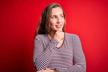 Obraz premium Young beautiful blonde woman wearing casual striped t-shirt over isolated red background with hand on chin thinking about question, pensive expression. Smiling with thoughtful face. Doubt concept.