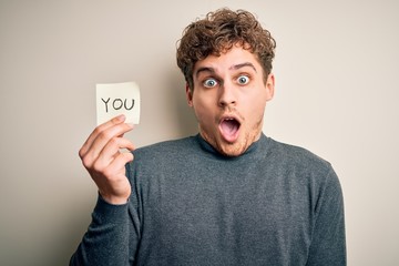 Young blond man with curly hair holding paper with you message over white background scared in shock with a surprise face, afraid and excited with fear expression