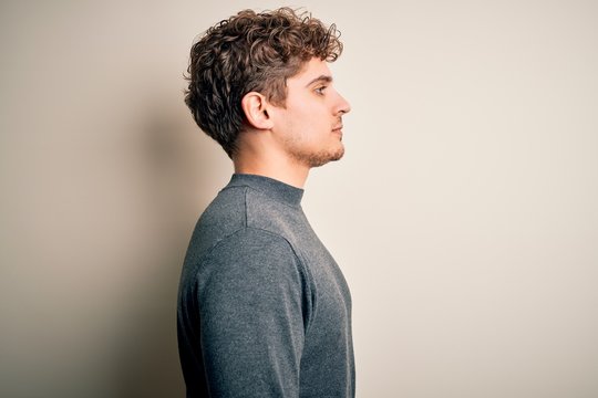 Young blond handsome man with curly hair wearing casual sweater over white background looking to side, relax profile pose with natural face with confident smile.