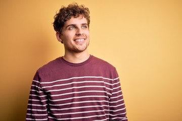 Young blond handsome man with curly hair wearing casual striped sweater looking away to side with smile on face, natural expression. Laughing confident.