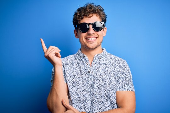 Young blond handsome man with curly hair on vacation wearing striped polo and sunglasses with a big smile on face, pointing with hand and finger to the side looking at the camera.