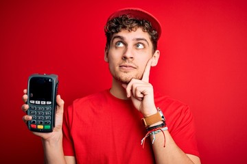 Young blond man with curly hair wearing cap holding dataphone over red background serious face...