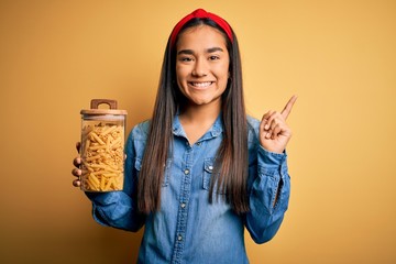 Young beautiful asian woman holding jar of Italian dry pasta macaroni over yellow background very happy pointing with hand and finger to the side