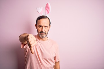 Middle age hoary man wearing bunny ears standing over isolated pink background looking unhappy and angry showing rejection and negative with thumbs down gesture. Bad expression.