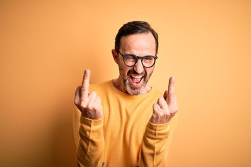 Middle age hoary man wearing casual sweater and glasses over isolated yellow background Showing middle finger doing fuck you bad expression, provocation and rude attitude. Screaming excited