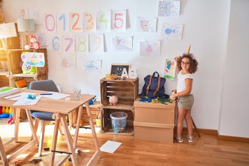 Beautiful toddler wearing glasses and unicorn diadem hanging up draw on the wall at kindergarten