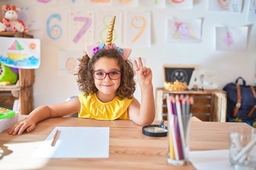 Beautiful toddler wearing glasses and unicorn diadem sitting on desk at kindergarten smiling with...