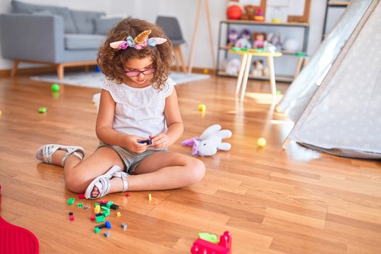 Beautiful toddler wearing glasses and unicorn diadem sitting on the floor playing with building blocks at kindergarten