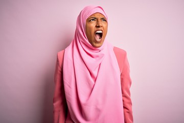 Young African American afro woman wearing muslim hijab over isolated pink background angry and mad screaming frustrated and furious, shouting with anger. Rage and aggressive concept.