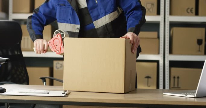Close up of hands of Caucasian postal male worker in uniform packing carton box at delievery department in post office. Postman closing parcel with sticky tape. Mail shipping concept.