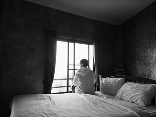 Back view of young man sitting alone on bed and looking through the window in bedroom