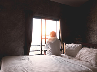 Back view of young man sitting alone on bed and looking through the window in bedroom