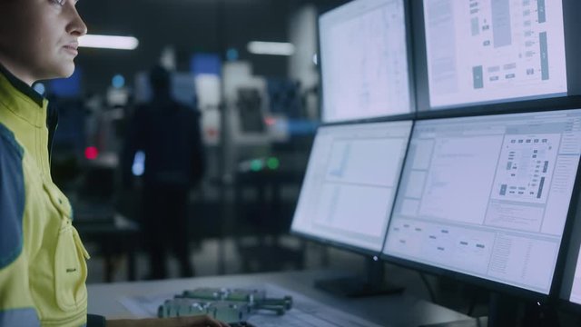 Industry 4.0 Modern Factory: Female Facility Operator Controls Workshop Production Line, Uses Computer with Screens Showing Complex UI of Machine Operation Processes, Controllers, Machinery Blueprints