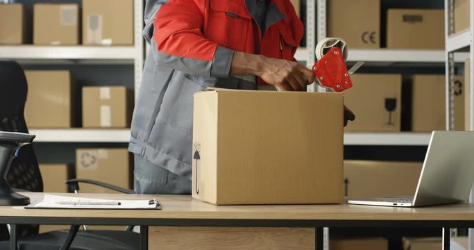 African American postal male worker in uniform packing carton box at delievery department in post office. Postman closing parcel with sticky tape. Mail shipping concept.