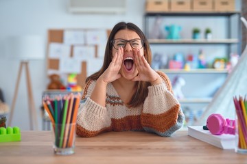 Young beautiful teacher woman wearing sweater and glasses sitting on desk at kindergarten Shouting angry out loud with hands over mouth