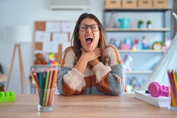 Young beautiful teacher woman wearing sweater and glasses sitting on desk at kindergarten shouting suffocate because painful strangle. Health problem. Asphyxiate and suicide concept.