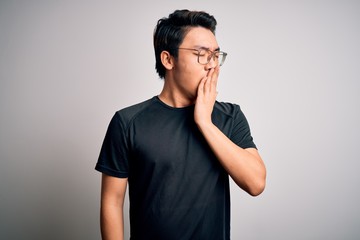 Young handsome chinese man wearing black t-shirt and glasses over white background bored yawning tired covering mouth with hand. Restless and sleepiness.