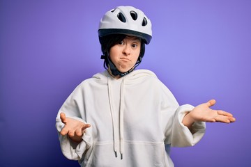 Young down syndrome cyclist woman wearing security bike helmet over purple background clueless and confused expression with arms and hands raised. Doubt concept.