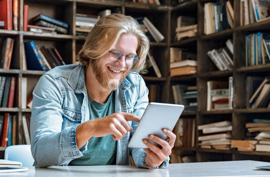 Young bearded man student entrepreneur hipster teacher glasses office home library desk hold modern tablet computer use app surf net social network watch video call smile tap touch screen copy space.