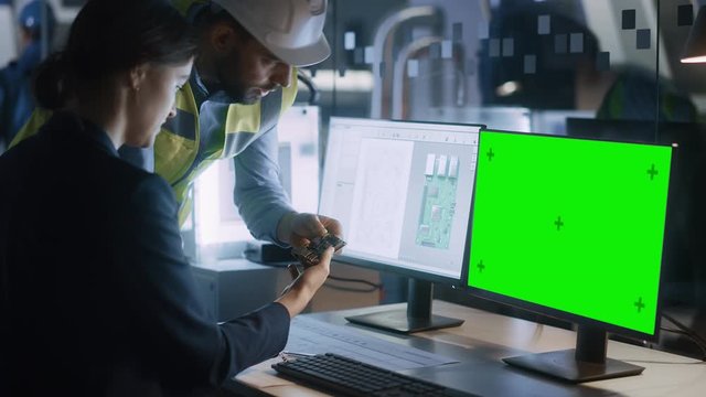 Supervisor Talks to Female Engineer Who's Working on a Computer, Two Monitor Screens Show Chroma Key, Green Screen Display and Processor Chip Design. Industry 4.0 Modern Electronics Production Factory