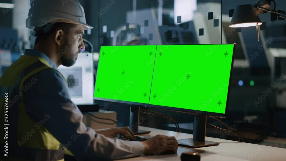 Poster engineer wearing safety vest and hardhat works on a computer, two monitor screens show chroma key / 