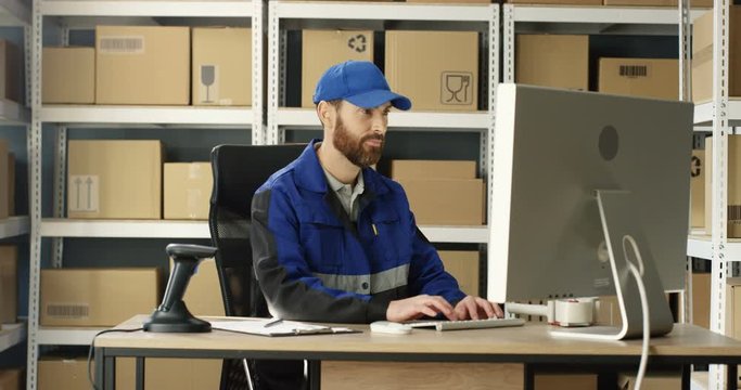 Handsome Caucasian mailman in blue iniform and cap sitting at table in postal office store and working at computer. Male post courier filling in invoice and entering data online in parcel storage.