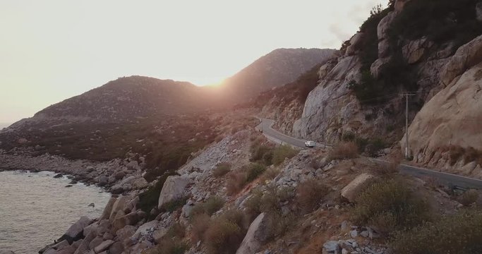 white car driving into the sunset on a bendy scenic road carved in a cliff next to the sea. Phn Rhang, Vietnam