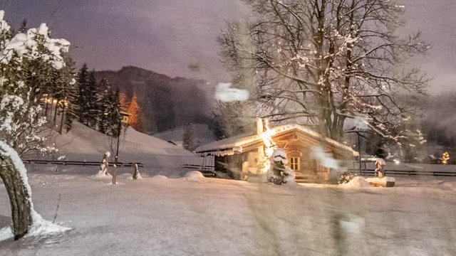 Winter fairy-tale scene. A small cottage decorated with bright, festive lights on the snow-covered field. Trees framing the scene, thick fluffs of snow laying on the branches.