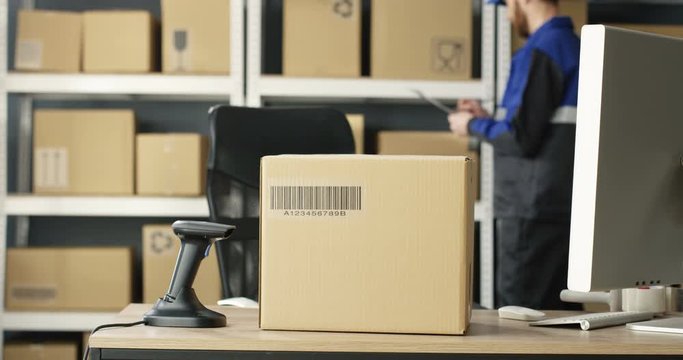 Close up of carton box with bar code is on table at computer in postal office store with parcels. Mail Caucasian courier with invoice and document working on background. Mailman counting boxes.