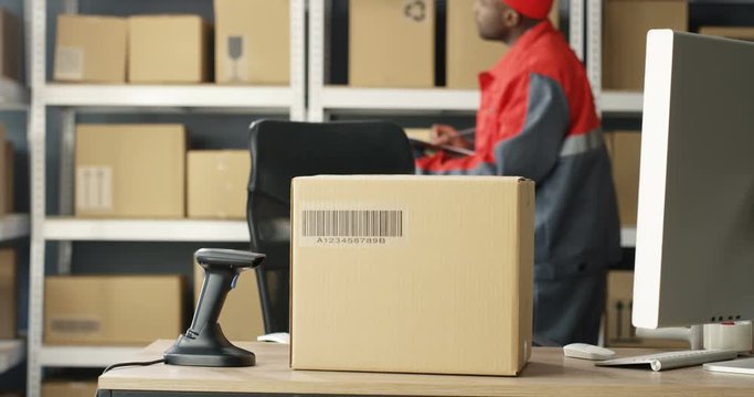 Close up of carton box with bar code is on table at computer in postal office store with parcels. Mail African American courier with invoice and document working on background. Mailman counting boxes.