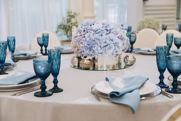 festive tables in the banquet room are decorated with compositions of white and blue flowers, on the tables are plates with napkins, glasses, candles, a wedding dinner