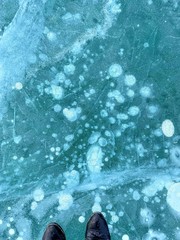 A view looking down at a pair of feet standing on frozen ice of a lake with interesting bubbles and...